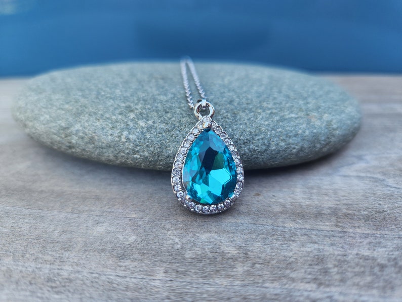 Puede incluir: Un collar con un colgante en forma de l&aacute;grima adornado con una gran piedra preciosa azul brillante rodeada por un halo de peque&ntilde;as piedras preciosas transparentes. El colgante es plateado y cuelga de una delicada cadena de plata.