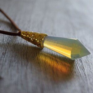 May include: A gold and clear crystal pendant necklace with a brown leather cord. The pendant is shaped like a long, pointed crystal.