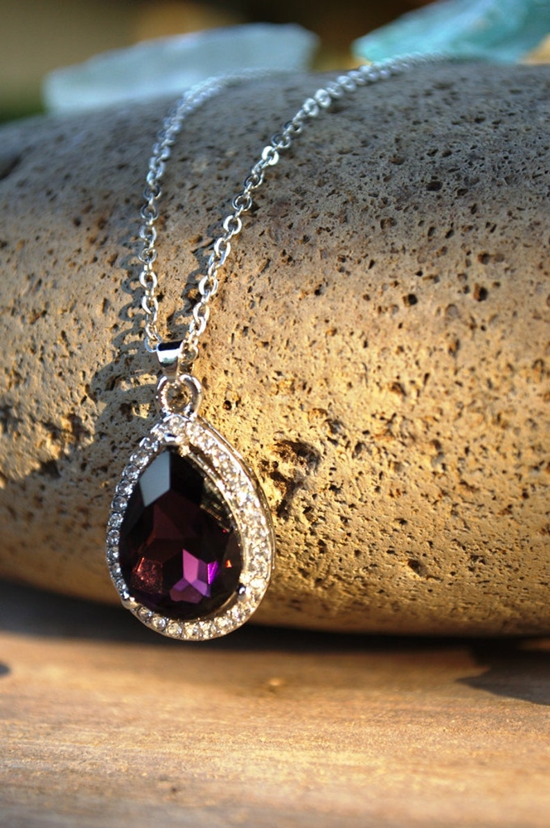 Puede incluir: Un collar de cadena de plata con un colgante en forma de l&aacute;grima que presenta una gran piedra preciosa de color p&uacute;rpura oscuro rodeada por una fila de piedras preciosas m&aacute;s peque&ntilde;as y transparentes.