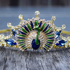 May include: A gold crown with a peacock design. The peacock is made of blue, green, and gold enamel. The crown is decorated with clear and blue rhinestones.