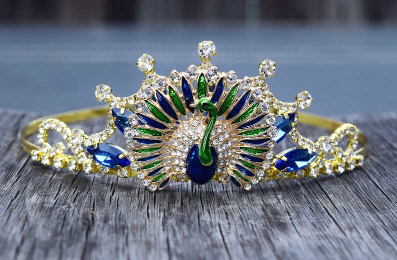 May include: A gold crown with a peacock design. The peacock is made of blue, green, and gold enamel. The crown is decorated with clear and blue rhinestones.