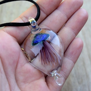 Pu&ograve; includere: Una collana con un ciondolo di vetro che contiene una medusa blu e viola. Il ciondolo ha la forma di una lacrima ed &egrave; sospeso da un cordoncino nero.