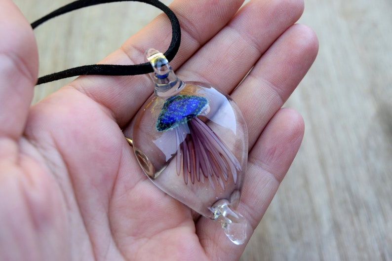 Pu&ograve; includere: Una collana con un ciondolo di vetro che contiene una medusa blu e viola. Il ciondolo ha la forma di una lacrima ed &egrave; sospeso da un cordoncino nero.