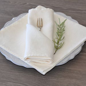 Può includere: Un tovagliolo di lino bianco piegato a forma di triangolo con una forchetta d'argento e un rametto di rosmarino in cima. Il tovagliolo è posto su un piatto bianco con bordo in pizzo.