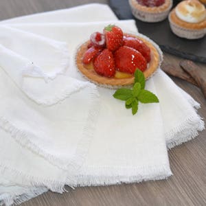 Può includere: Un tovagliolo di lino bianco con un bordo a frangia, piegato e posizionato su un tavolo di legno. Una piccola torta con ripieno di fragole è sopra il tovagliolo. Un rametto di menta fresca è accanto alla torta.