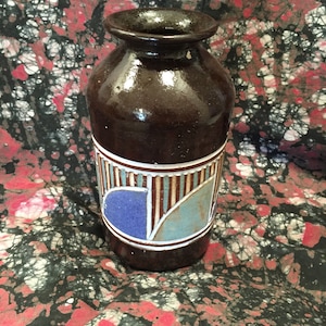May include: A brown ceramic vase with a decorative band featuring blue, white, and tan geometric shapes. The vase has a rounded body and a flared rim. The background is a fabric with red, black, and white patterns.