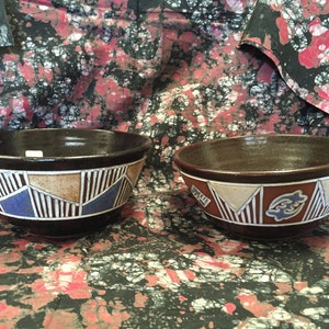 May include: Two handmade ceramic bowls. The bowls are dark brown with a lighter brown interior. The exterior features a geometric design with blue, tan, and white accents. The bowls are displayed on a patterned fabric background.