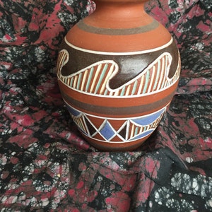 May include: A terracotta ceramic vase with a brown rim and base. The vase has horizontal bands of brown, white, and blue geometric designs. The vase has a rounded body and a narrow neck. The background is a patterned fabric with red and black hues.