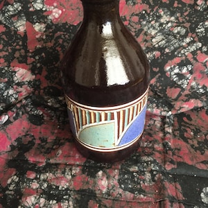 May include: A dark brown ceramic bottle with a glossy finish. The bottle features a decorative band with geometric shapes in blue, teal, and white. The bottle has a narrow neck and a rounded body. The background is a patterned fabric.