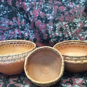 May include: Three light brown pottery bowls with woven rims. The bowls are of varying sizes, with the largest on the right and the smallest in the centre. The background features a patterned fabric with red and black hues.
