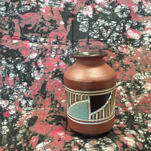 May include: A brown ceramic vase with a cylindrical body and a narrow neck. The vase features a geometric design with vertical lines, black, and teal accents. The background is a fabric with a black, white, and red pattern.