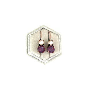 May include: A pair of dangle earrings with a small white pearl and a larger purple gemstone. The earrings are on a white velvet background.