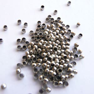 May include: A scattering of small, shiny, silver-coloured metal beads on a white surface. The beads are round with a hole through the centre, and vary slightly in size. These beads are likely used for jewellery making or other crafts.