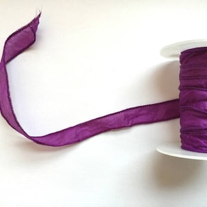May include: A spool of purple satin ribbon with a slightly textured surface. The ribbon is wound around a white plastic spool.