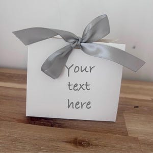 May include: A white gift box tied with a grey satin ribbon bow. The box has the words "Your text here" printed on the front in a grey font. The box is on a wooden surface.