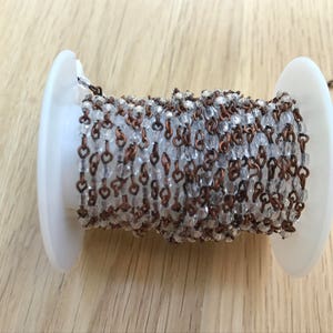 May include: A spool of decorative chain featuring clear, faceted beads and copper-colored links. The chain is wound around a white plastic spool, with a small length extending from the side. Ideal for jewelry making or craft projects.