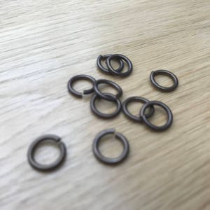 May include: A selection of small, dark grey metal split rings scattered on a light wood surface. The rings are open, and some are interlocked. The image is a close-up, showing the texture of the rings and the wood grain.