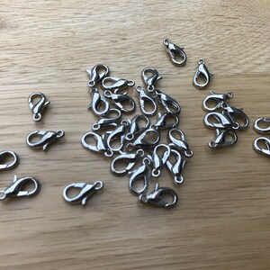 May include: A selection of silver lobster claw clasps, scattered on a light brown wooden surface. These clasps are used in jewellery making and crafting to secure necklaces, bracelets, and other accessories.