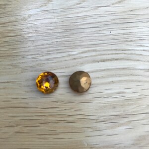May include: Two small, round, decorative objects on a light wood surface. One is a gold-coloured metal button and the other is a clear, faceted, amber-coloured glass button.