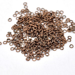 May include: A collection of small, bronze-coloured jump rings scattered on a white surface. These open metal rings are used for jewellery making and other crafts. The rings are circular and have a split, allowing them to be opened and closed.