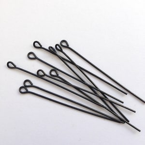 May include: A selection of black eye pins, each with a looped end, scattered on a white surface. These pins are slender and straight, suitable for jewellery making and crafting projects. The eye pins are made of metal.