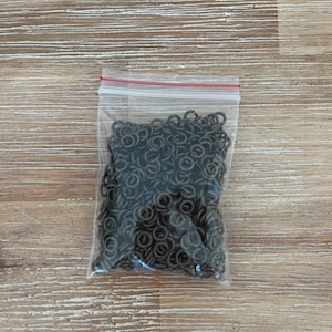 May include: A clear plastic bag filled with a large quantity of small, dark brown metal jump rings.
