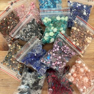 May include: Assortment of 11 small plastic bags filled with various colourful beads. The bags are labelled with numbers 1 through 11. The beads are in a variety of shapes, sizes, and colours, including clear, pink, blue, green, red, and brown.