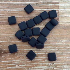 May include: A selection of matte black square beads, each with rounded edges and two holes, scattered on a light brown wooden surface. Suitable for jewellery making and crafts.