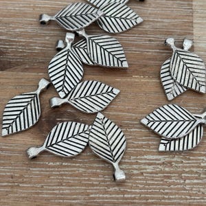 May include: A collection of silver-colored leaf charms with detailed veining. Each charm has a loop at the top for easy attachment. The charms are scattered on a wooden surface, ideal for jewelry making or craft projects.