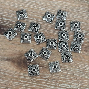 May include: Silver square beads with an ornate design. Each bead has a central hole surrounded by a circular pattern and decorative swirls. The beads are scattered on a wooden surface.