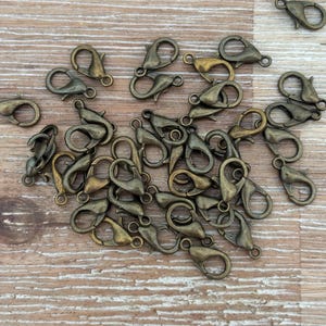 May include: A selection of antique bronze-coloured lobster claw clasps, arranged on a light brown wooden surface. The clasps are small, with a curved design and a spring-loaded mechanism for secure closure. Suitable for jewellery making and crafting projects.