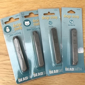 May include: Four design stamps in packaging, each with a different 6mm design: ladybug, cartoon cat, vine, and baby feet. The stamps are silver-coloured and are labelled "BEADSMITH". The packaging is light blue.