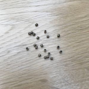 May include: A scattering of small, cylindrical, dark gray metal beads. These beads are evenly spaced across a light-colored wooden surface. The beads appear to be hollow and have a small hole running through the center.