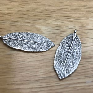 May include: Two silver leaf-shaped charms with detailed etched designs. The charms have a loop at the top for attaching to jewellery or other crafts.