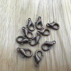 May include: A collection of bronze-colored lobster claw clasps. These small metal clasps are designed for jewelry making and crafting. The clasps are arranged on a light brown wooden surface.