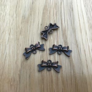 May include: Four bronze-colored metal bow charms with a distressed finish, each featuring a bow design with a loop for attaching to jewelry or crafts. The charms are arranged on a light wood-grain surface.