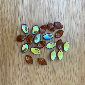 May include: A scattering of amber-hued, leaf-shaped glass beads with iridescent finishes. The beads are arranged on a light brown wooden surface, highlighting their diverse forms and reflective properties. These decorative beads are ideal for jewellery making or craft projects.