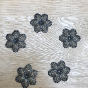 May include: Five silver metal flower-shaped embellishments with intricate designs. The flowers are arranged in a circular pattern on a light wood surface.