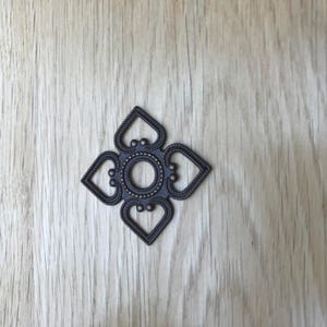 May include: A dark bronze-colored decorative metal applique in a symmetrical floral design. The applique features a central circle surrounded by four heart-shaped elements, each with a decorative border. The piece is set against a light wood grain background.