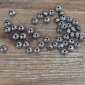 May include: A scattering of small, silver-coloured beads. Each bead features a detailed, ridged design with a central hole and a decorative pattern. The beads are arranged on a light brown wooden surface.