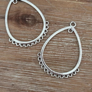 May include: Two silver-coloured teardrop-shaped earring components. Each component has a loop at the top for attachment and a scalloped edge along the bottom. The components are displayed on a wooden surface.