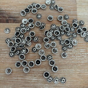 May include: A collection of small, silver-colored bead caps scattered on a light brown wooden surface. Each cap has a decorative, scalloped edge and a central hole, ideal for jewelry making or crafting projects.