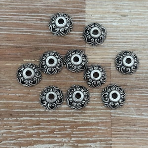 May include: Ten silver-coloured decorative beads are displayed. Each bead has a floral design with heart-shaped details and a central hole. They are arranged on a light brown wooden surface.