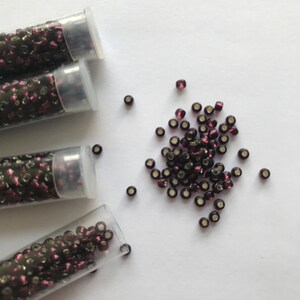 May include: Four small clear tubes filled with dark purple seed beads. The beads are scattered on a white surface.