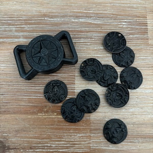 May include: Black compass buckle and several black decorative coins with a detailed relief design. The buckle features a compass rose design. The coins have a detailed relief design. The items are on a light brown wooden surface.