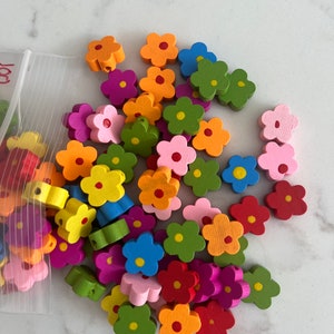 May include: A selection of brightly coloured wooden flower beads in shades of pink, orange, green, blue, yellow, and red. Each bead has a small hole for threading. The beads are scattered on a white surface, with some in a clear plastic bag.