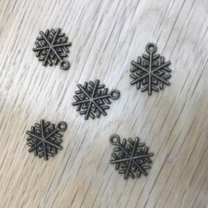 May include: Five bronze snowflake charms, each with a loop for hanging. They are displayed on a light brown wooden surface. These decorative charms are suitable for jewellery making or festive crafts.