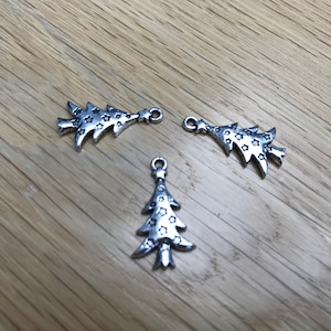 May include: Three silver-colored Christmas tree charms with star cutouts. Two charms are stylized, and one is a traditional tree shape. Each charm has a small loop for attaching to jewelry or other crafts. The charms are on a light brown wooden surface.