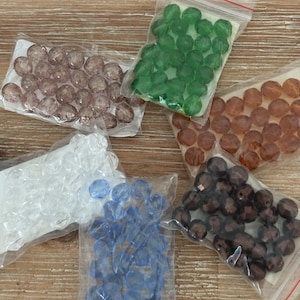 May include: Six small bags of assorted glass beads. The bags contain clear, light blue, dark blue, brown, amber, and green beads.
