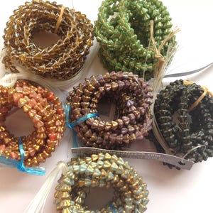 May include: Assortment of glass beads in various colours, including brown, green, pink, purple, and blue. The beads are all in the shape of a cone and have a metallic finish. The beads are arranged in a circle on a white surface.